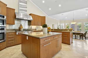 Kitchen with wall chimney range hood, a sink, a center island, stainless steel appliances, and light stone countertops