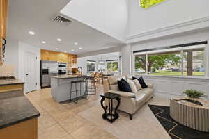 Open concept kitchen, living room, and dining space, featuring large windows with custom roller shades, mixed tile and patterned carpet flooring