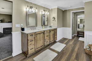 Bathroom with a fireplace, wood finished floors, ornamental molding, double vanity, and wainscoting