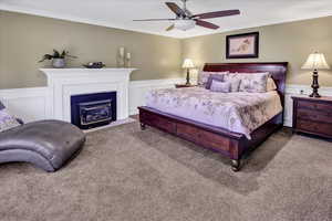 Carpeted bedroom with crown molding, wainscoting, a decorative wall, a brick fireplace, and a ceiling fan