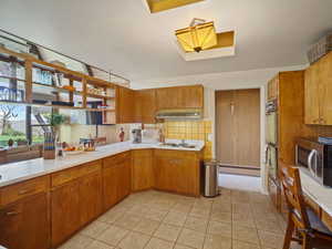 Kitchen featuring stainless steel microwave, under cabinet range hood, light countertops, brown cabinets, and a skylight