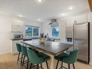 Kitchen featuring stainless steel refrigerator with ice dispenser, dark countertops, wall chimney exhaust hood, a kitchen bar, and white cabinetry