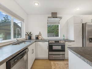 Kitchen with stainless steel appliances, wall chimney exhaust hood, a sink, light wood-style floors, and white cabinets