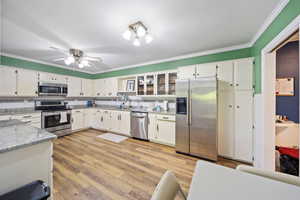 Kitchen with stainless steel appliances, crown molding, white cabinets, light stone counters, and light wood-style floors