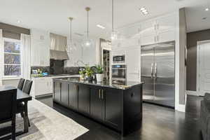 Kitchen featuring built in appliances, wall chimney exhaust hood, white cabinets, dark cabinets, and dark stone counters