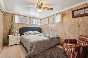Bedroom with light carpet, a textured ceiling, wooden walls, crown molding, and ceiling fan