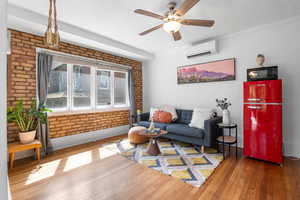 Living area featuring a wall mounted air conditioner, hardwood / wood-style floors, a ceiling fan, brick wall, and baseboards