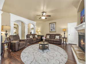 Living area with decorative columns, a ceiling fan, a tiled fireplace, arched walkways, and dark wood finished floors