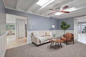 Living area featuring healthy amount of natural light, beam ceiling, a ceiling fan, a wainscoted wall, and light carpet