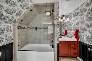 Full bath featuring wallpapered walls, a wainscoted wall, shower / bath combination with glass door, and vanity
