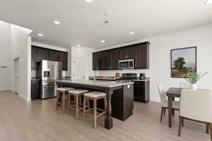 Kitchen featuring stainless steel appliances, light wood-type flooring, a kitchen island with sink, a breakfast bar, and dark brown cabinetry