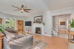 Living area featuring stairs, ornamental molding, ceiling fan, a lit fireplace, and light wood-style floors