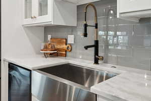 Kitchen view of backsplash, white cabinetry, glass insert cabinets, light stone counters, and a stainless-steel sink