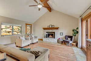 Living room featuring light wood-style flooring, ceiling fan, beam ceiling, a stone fireplace, and high vaulted ceiling