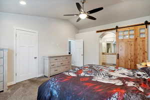 Carpeted bedroom with vaulted ceiling, a barn door, ensuite bath, baseboards, and ceiling fan