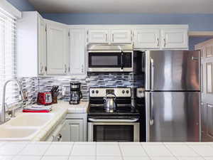 Kitchen with appliances with stainless steel finishes, tile countertops, a sink, white cabinets, and tasteful backsplash