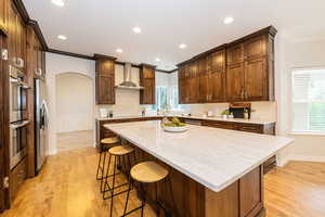 Kitchen with wall chimney exhaust hood, crown molding, arched walkways, stainless steel appliances, and light wood-style flooring