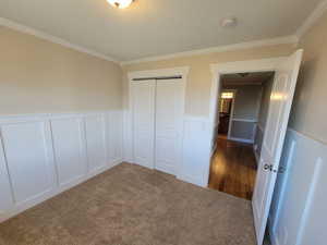 Unfurnished bedroom featuring a decorative wall, ornamental molding, wainscoting, a closet, and dark colored carpet