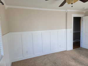 Unfurnished bedroom with a decorative wall, ornamental molding, carpet floors, a wainscoted wall, and a ceiling fan