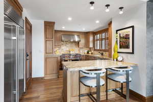 Kitchen with high quality appliances, wall chimney range hood, light wood finished floors, a peninsula, and recessed lighting