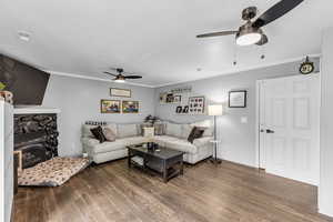 Living area with ceiling fan, wood finished floors, ornamental molding, and a stone fireplace