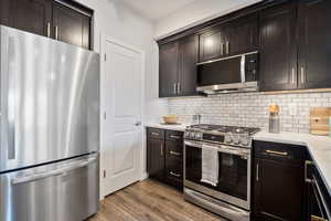 Kitchen with stainless steel appliances, decorative backsplash, wood finished floors, dark brown cabinetry, and light stone countertops