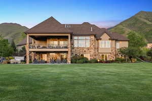 Back of house at dusk featuring a lawn, stone siding, a shingled roof, and a mountain view