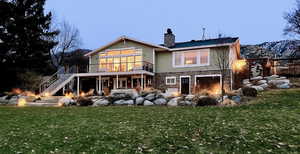 Back of house at dusk featuring stairs, stone siding, a chimney, a yard, and a wooden deck