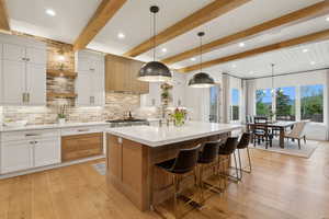 Kitchen featuring light countertops, light wood finished floors, decorative backsplash, an island with sink, and beam ceiling