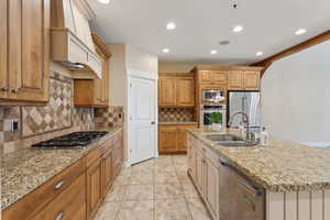 Kitchen with a sink, appliances with stainless steel finishes, a kitchen island with sink, tasteful backsplash, and custom range hood