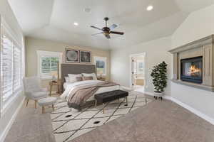 Carpeted bedroom featuring vaulted ceiling, a fireplace, baseboards, connected bathroom, and a ceiling fan