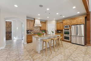 Kitchen with appliances with stainless steel finishes, custom exhaust hood, arched walkways, light stone countertops, and a sink