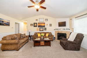 Living area with vaulted ceiling, a ceiling fan, wainscoting, light colored carpet, and a tiled fireplace