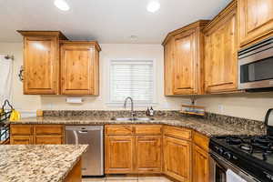 Kitchen with stainless steel appliances, a sink, dark stone counters, brown cabinetry, and recessed lighting