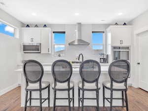 Kitchen featuring stainless steel appliances, wall chimney range hood, a center island with sink, decorative backsplash, and white cabinetry