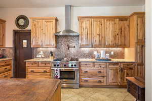 Kitchen with stainless steel range, wall chimney exhaust hood, backsplash, brown cabinets, and light stone countertops
