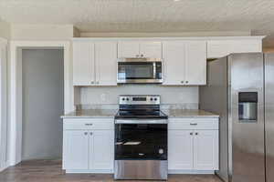 Kitchen with stainless steel appliances, wood finished floors, white cabinetry, a textured ceiling, and light stone counters