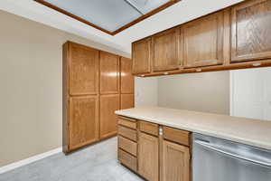 Kitchen featuring stainless steel dishwasher, light countertops, brown cabinetry, and baseboards