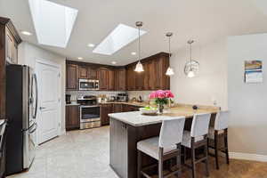 Kitchen with appliances with stainless steel finishes, a peninsula, a skylight, dark brown cabinetry, and a breakfast bar area