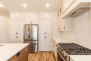 Kitchen with stainless steel appliances, light wood-style flooring, custom exhaust hood, white cabinetry, and glass insert cabinets