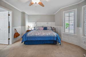 Bedroom with crown molding, carpet, a wainscoted wall, a ceiling fan, and a decorative wall
