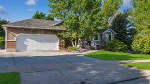 View of property hidden behind natural elements featuring driveway, a front yard, an attached garage, roof with shingles, and brick siding
