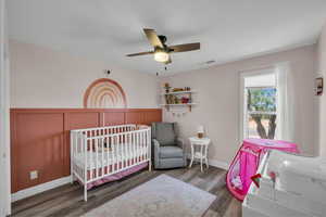 Bedroom featuring wood finished floors, a ceiling fan, wainscoting, a decorative wall, and baseboards