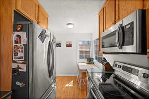 Kitchen featuring appliances with stainless steel finishes, light wood-style flooring, a textured ceiling, baseboards, and brown cabinets