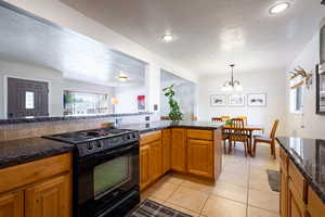 Kitchen with black electric range oven, a textured ceiling, a chandelier, wood cabinets, and dark granite counters