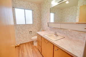 Bathroom featuring wood finished floors, wallpapered walls, toilet, vanity, and baseboards