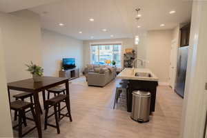 Kitchen with stainless steel fridge, a sink, light wood finished floors, a center island with sink, and recessed lighting