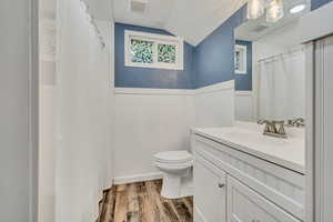 Full bath with vaulted ceiling, toilet, wood finished floors, wainscoting, and vanity