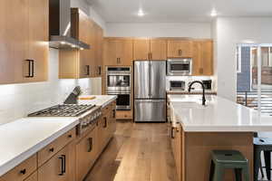 Kitchen featuring appliances with stainless steel finishes, wall chimney range hood, a sink, light wood-style floors, and light countertops