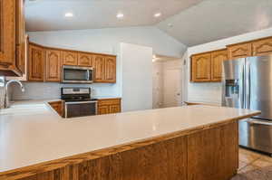 Kitchen with appliances with stainless steel finishes, a sink, vaulted ceiling, light countertops, and brown cabinetry